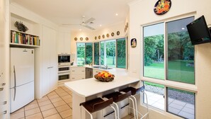 A modern kitchen with white cabinetry, a central island with stools, a microwave, and a fruit bowl. - Luxury Home in Nature surrounded by Wallabies (Clifton Beach)