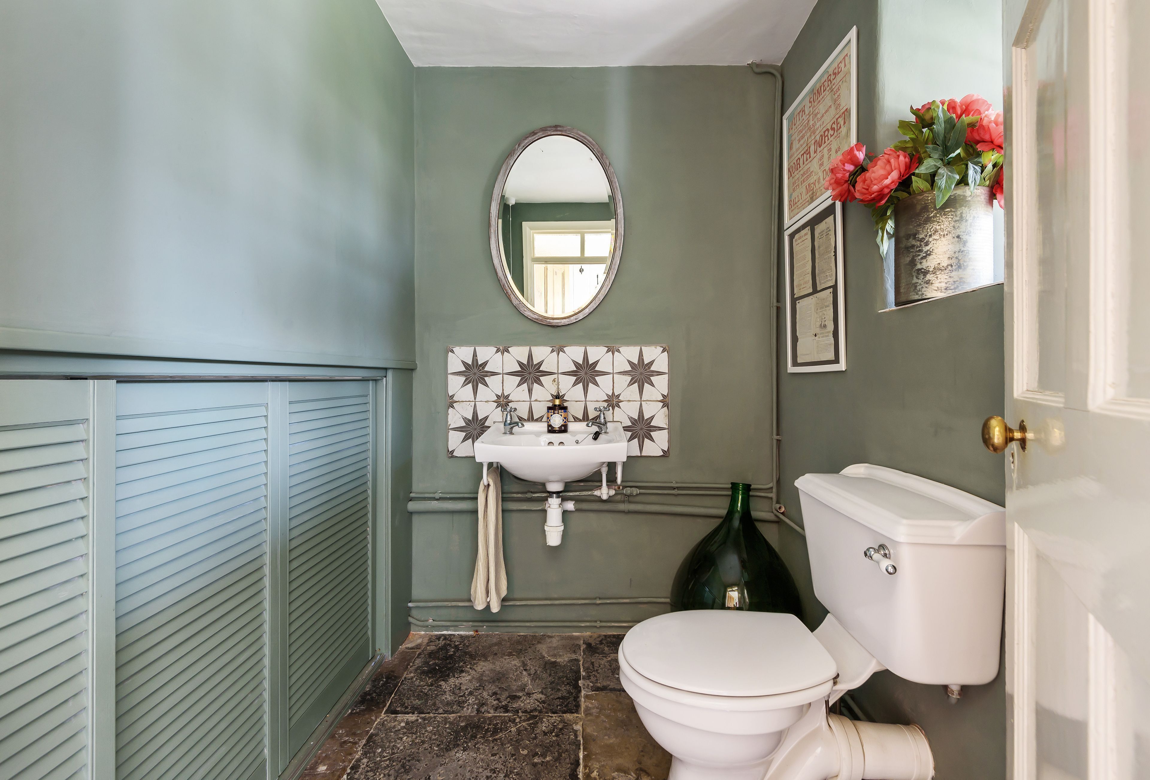 Et badeværelse med et hvidt toilet, en vask med et spejl over og en dekorativ vase med blomster på væggen.