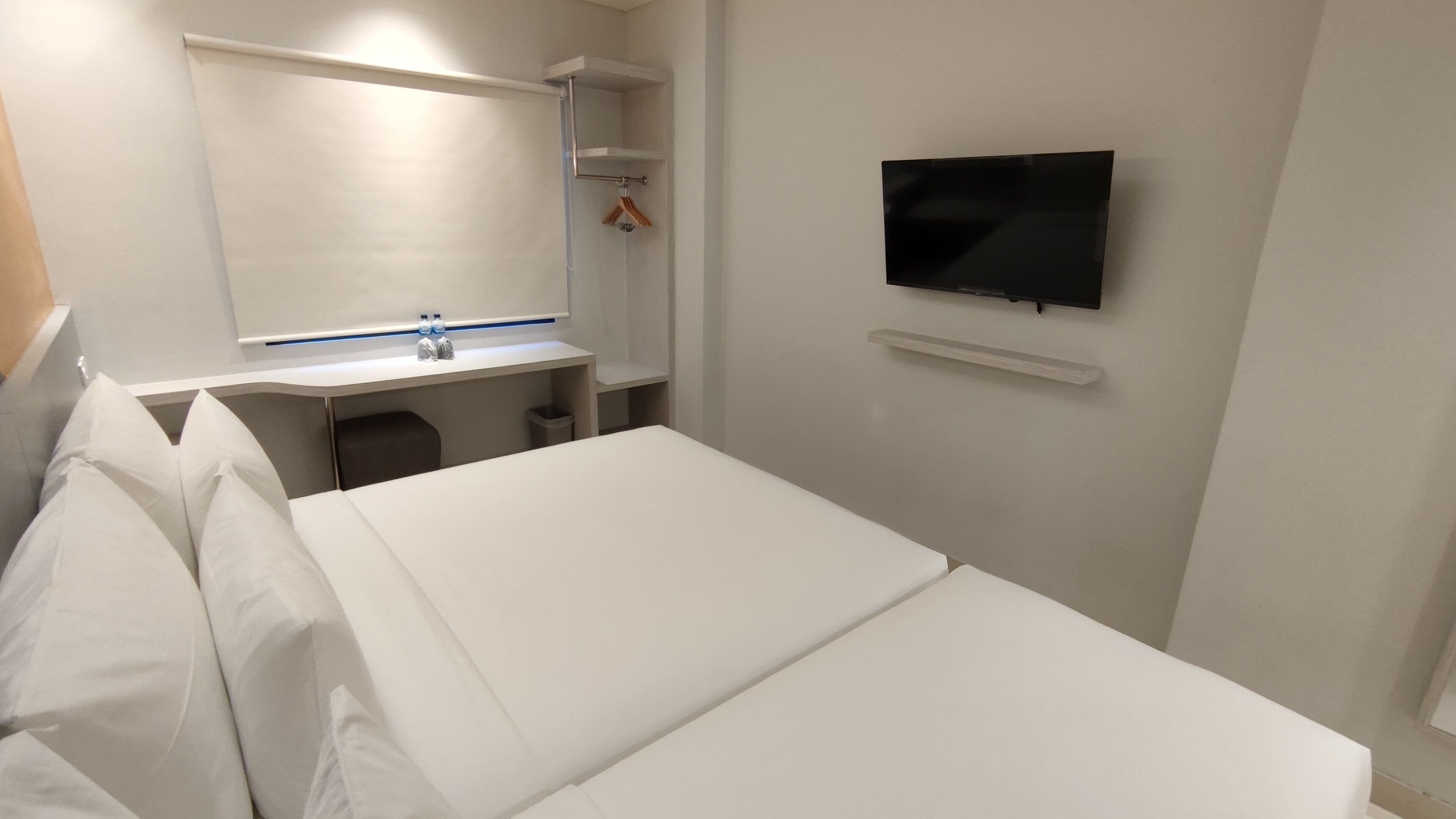 Ett hotellrum med en säng, ett skrivbord med en spegel, en tv som är monterad på väggen och en liten hylla.