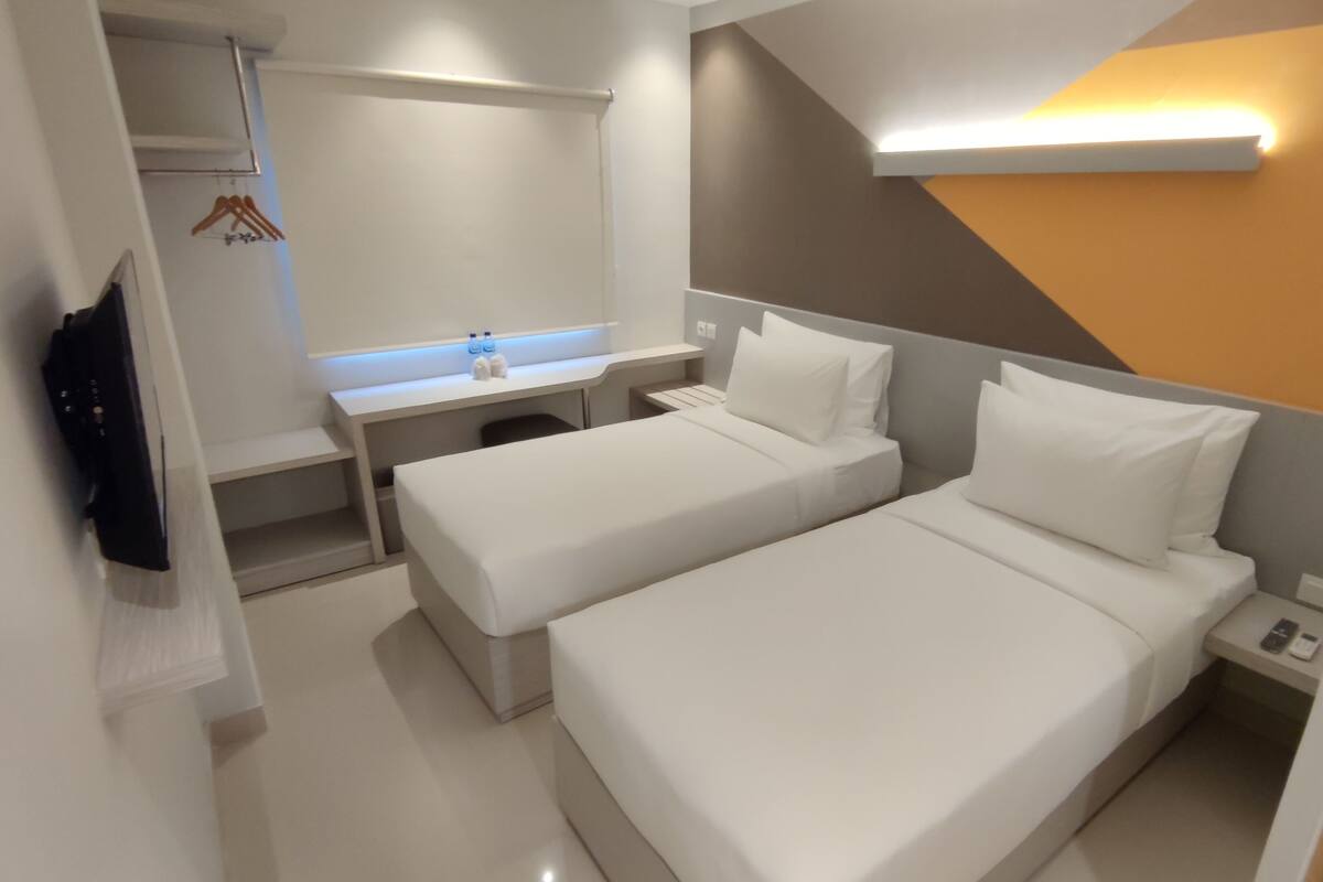A modern hotel room with two single beds, a desk with a chair, a TV, and a wardrobe.