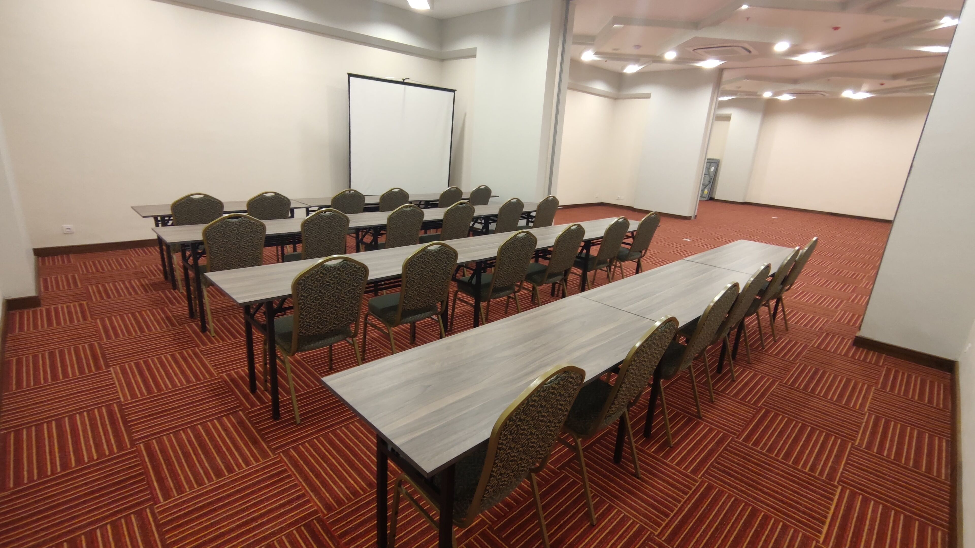 A conference room with multiple tables and chairs, carpeted floor, whiteboard, and a projector screen.