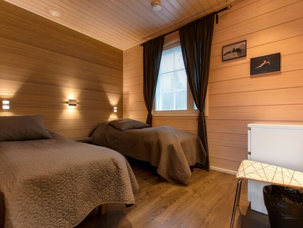 A cozy room with wooden panel walls, two beds with matching bedding, a window with curtains, and a small table with a trash bin.