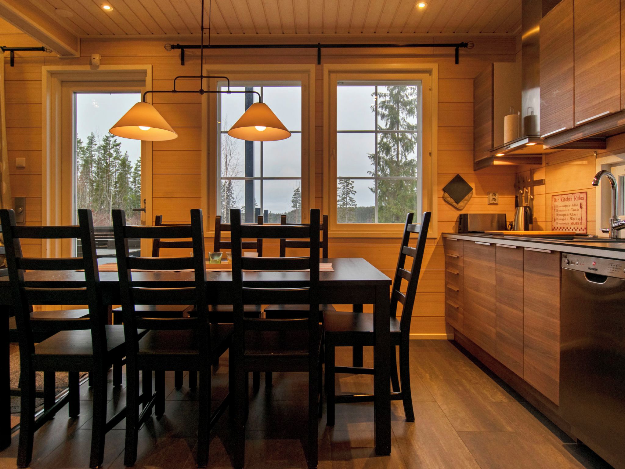 A wooden interior with a dining table and chairs, large windows, and a kitchen area.