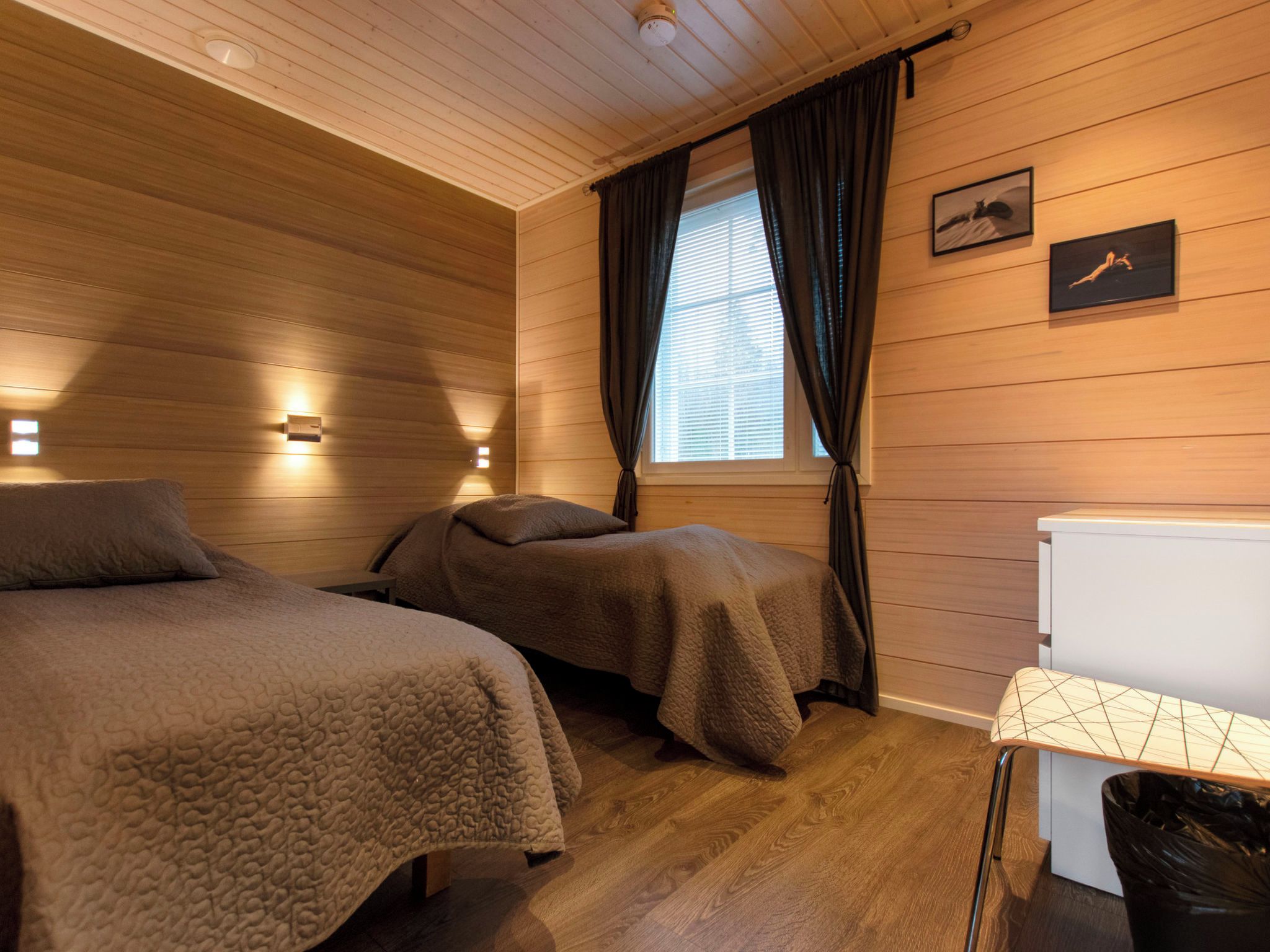 A room with two beds, a window with curtains, a wall-mounted light, and a small table with a trash bin.