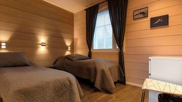 A room with two beds, a window with curtains, a wall-mounted light, and a small table with a trash bin.
