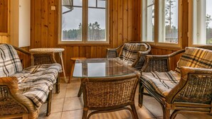 Una habitación acogedora con paredes de madera y ventanas amplias, amueblada con sillas de mimbre y una mesa de vidrio.