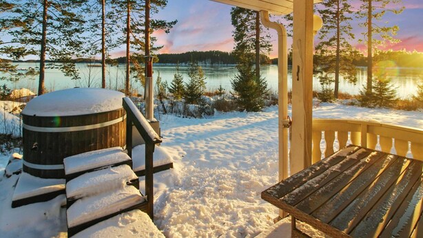 Una terraza de madera con un banco y un barril, con vistas a un lago con árboles y un atardecer.