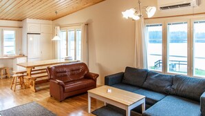 Een gezellige woonkamer met een bruine leren bank, een grijze bank en een houten salontafel. Er is een houten eettafel met stoelen, een keuken met witte kasten en grote ramen met uitzicht op het buitenlandschap.