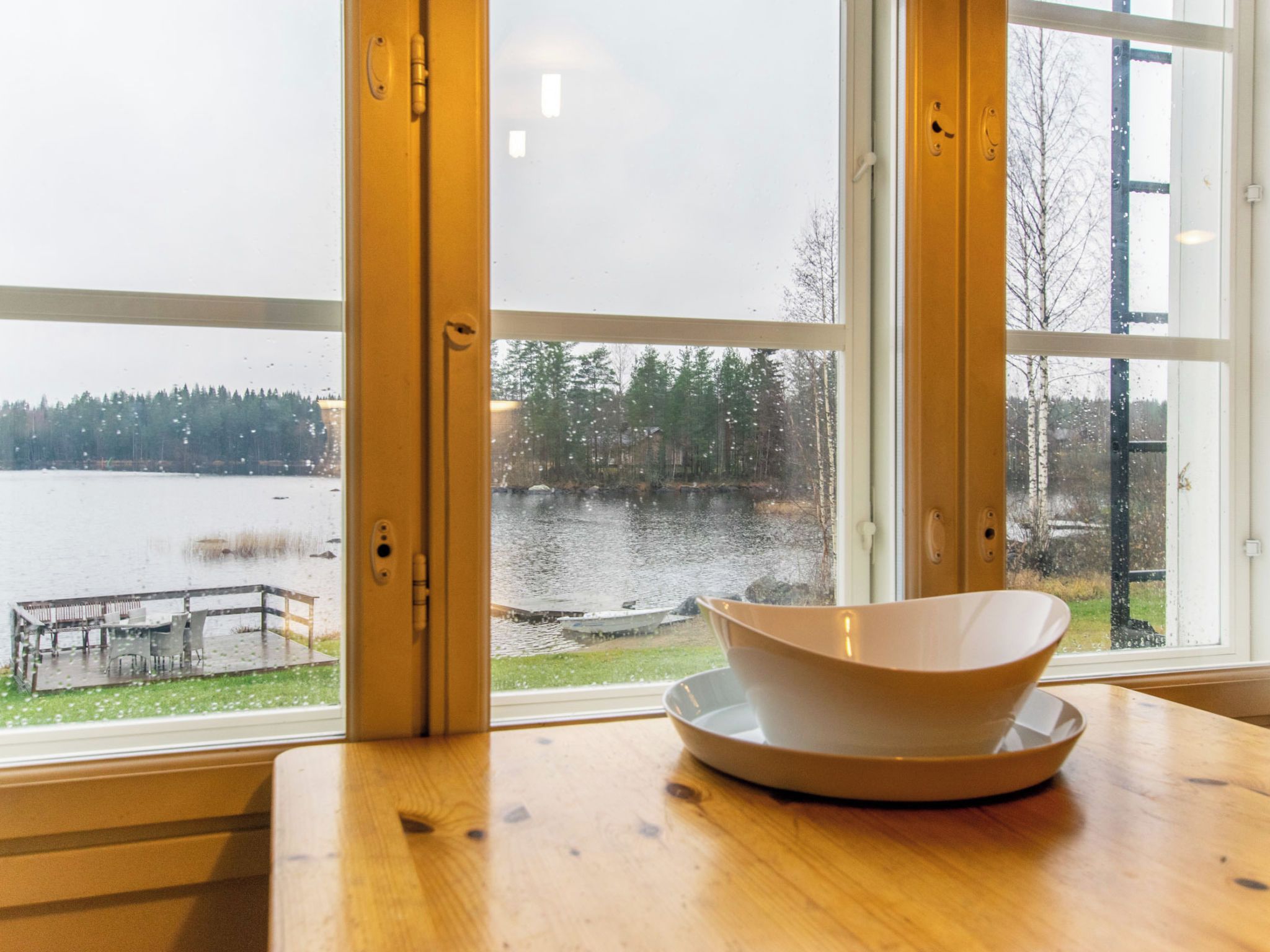 Een kamer met een groot raam met uitzicht op een meer, een houten tafel met een kom, en een bank buiten.