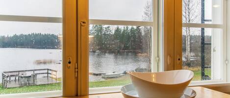 Una habitación con un ventanal grande que da a un lago, una mesa de madera con un cuenco, y una banca afuera.