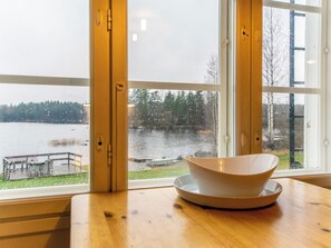 Una habitación con un ventanal grande que da a un lago, una mesa de madera con un cuenco, y una banca afuera.