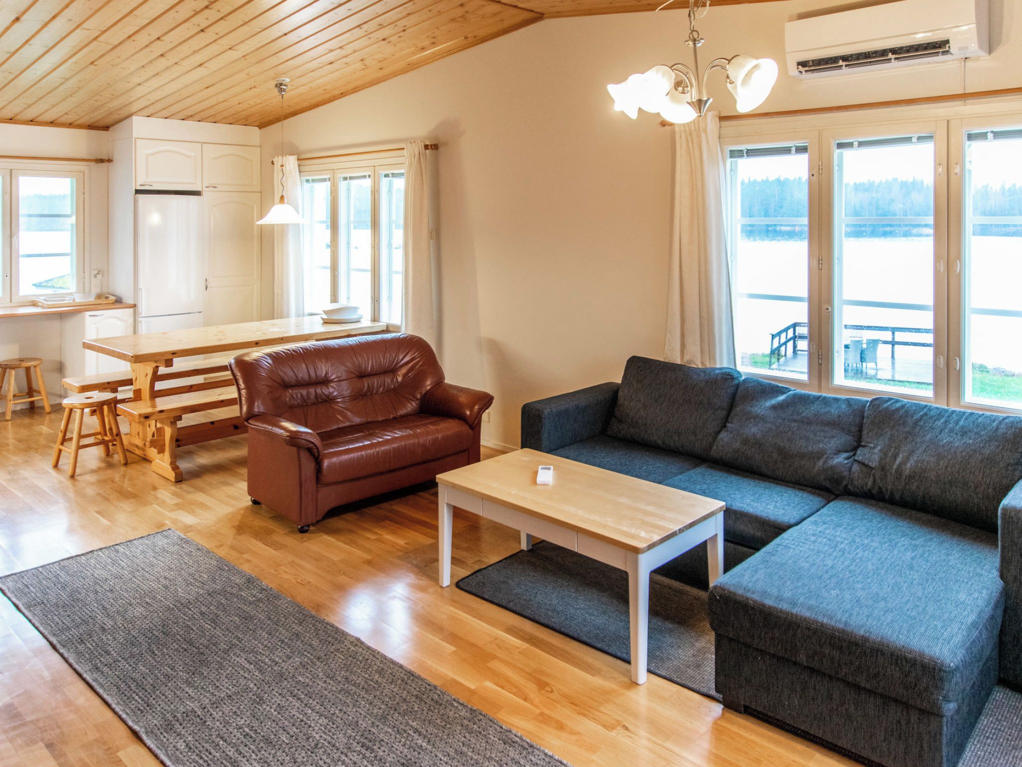 Ett rymligt vardagsrum med ett träpanelat tak, en brun lädersoffa, en grå soffa, ett träbord och ett kök i bakgrunden.