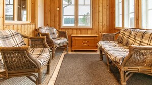 Een gezellige kamer met houten wanden en grote ramen, ingericht met rieten stoelen en een bank, en een houten dressoir.