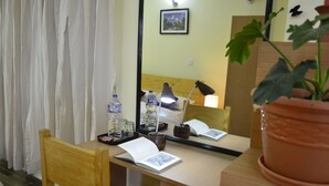 Quarto de hotel com escrivaninha de madeira, uma cadeira, planta em vaso, espelho, abajur e janela com cortinas.