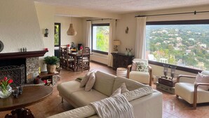 A spacious living room with a large window offering a view of a town. There is a fireplace, a dining table with chairs, and a sofa. - Villa Schenk - Urb Rio de Oro, Calonge, Catalonia (Calonge)