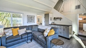 Een moderne woonkamer met een grijze hoekbank, gekleurde kussens en een kamerkleed met een geometrisch patroon. Een klein houten krukje en bijzettafeltje zijn zichtbaar. De kamer heeft een trap die leidt naar een keuken met witte kasten en een aanrecht.
