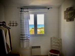 Ein Zimmer mit Blick auf einen Strand, ein weißes Stuhl, ein Kleiderständer mit Kleidung und eine Wandleuchte.