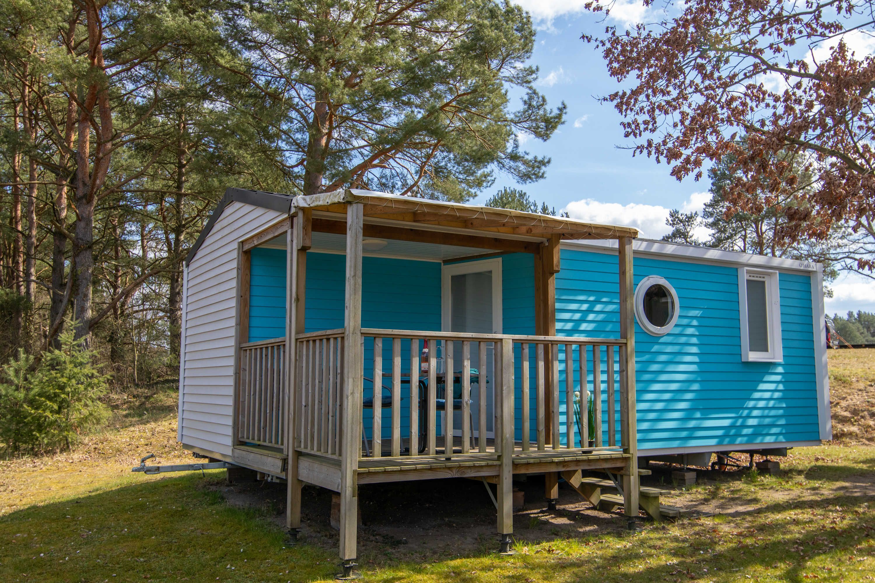 A small, single-story mobile home with a wooden deck and a porch, surrounded by trees.