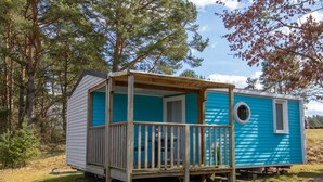 A small, single-story mobile home with a wooden deck and a porch, surrounded by trees.