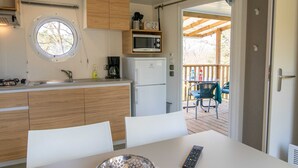 A compact kitchen with a microwave, coffee maker, and a window. There is a table with a remote control and a bowl. A door leads to an outdoor area with a deck and chairs.