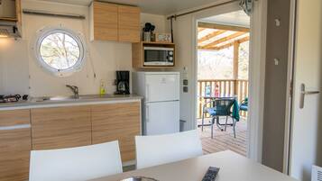 A compact kitchen with a microwave, coffee maker, and a window. There is a table with a remote control and a bowl. A door leads to an outdoor area with a deck and chairs.