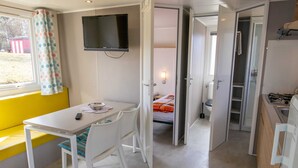 A compact room with a bed, a small table with a chair, a TV mounted on the wall, and a window with a curtain.