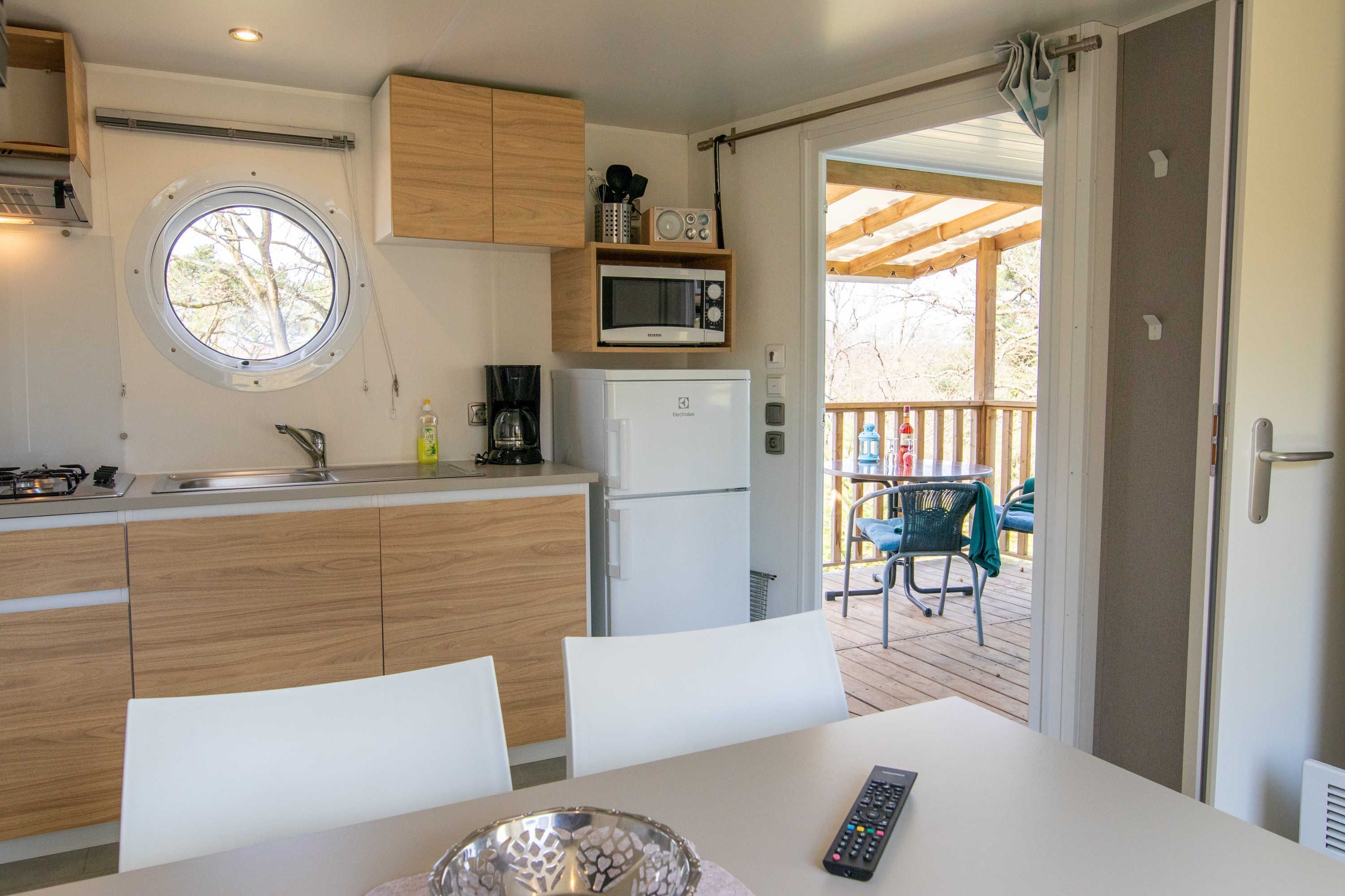 A compact kitchen with a microwave, sink, and wooden cabinets. There's a dining table with chairs and a remote control on it.
