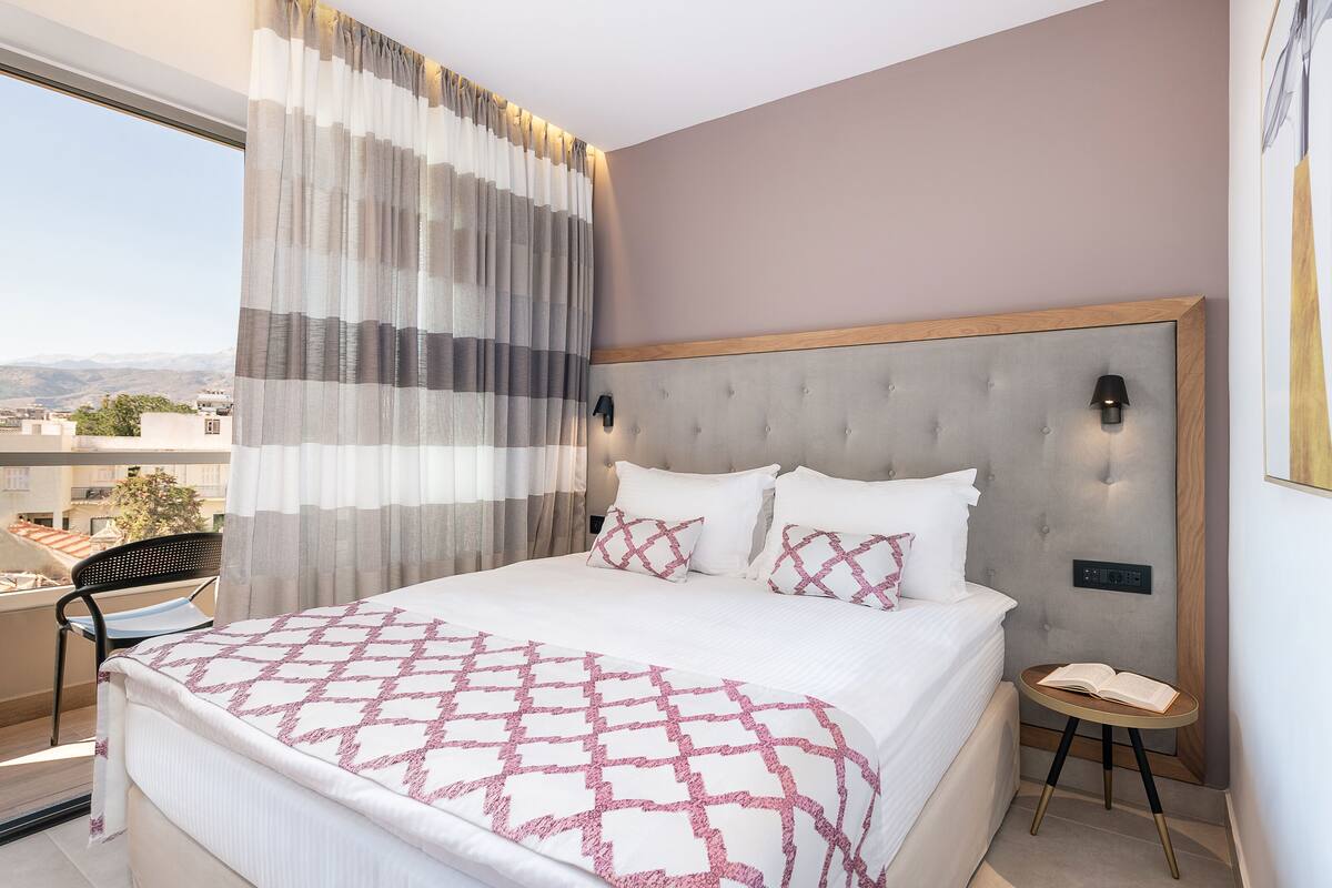 A hotel room with a large bed, a bedside table, a view of the city, and a balcony.