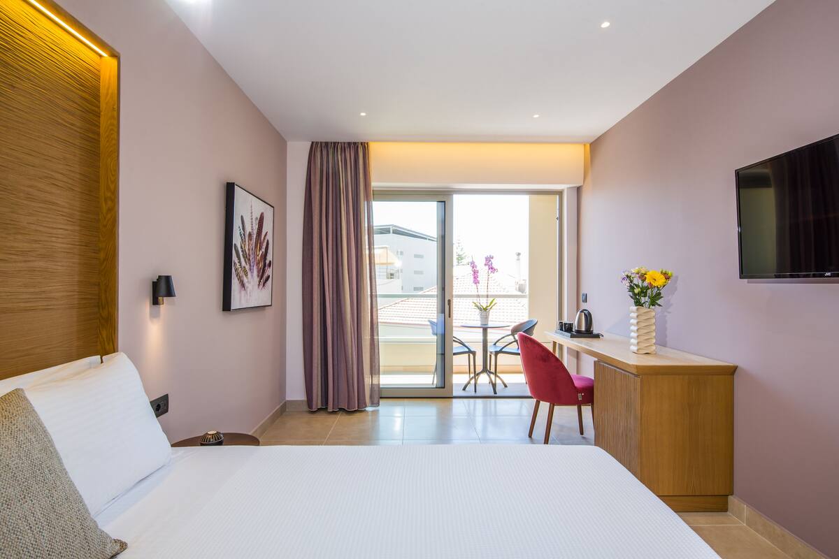 A hotel room with a bed, a desk with a chair, a TV, and a balcony with a view of buildings and flowers.