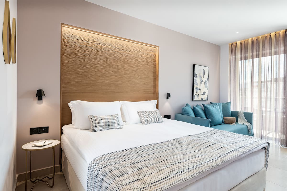 A modern hotel room with a large bed, a bedside table, a sofa, and a large window with curtains.