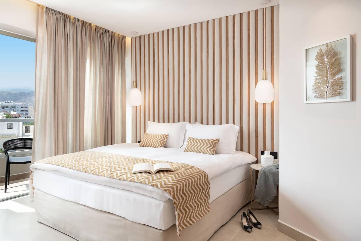 A modern hotel room with a large bed, striped wallpaper, a framed leaf artwork, and a view of the city.