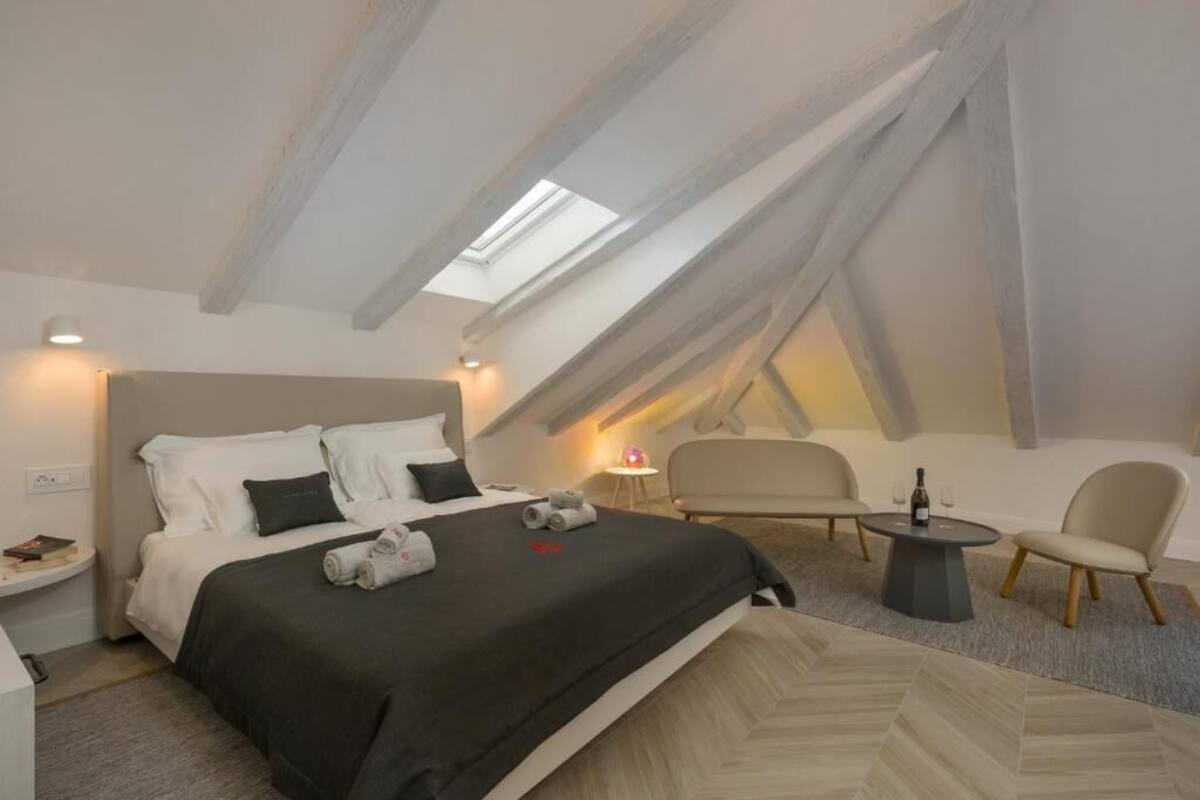 Un dormitorio con tragaluz, una cama con almohadas, una mesita de noche, una silla y una pequeña mesa redonda con una botella y vasos.