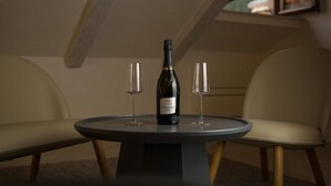 Mesa redonda con una botella de vino y dos copas, ubicada entre dos sillas en una habitación con techo inclinado y una ventana.