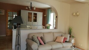 Un salon avec un canapé beige, une table basse en verre et un coin cuisine doté de placards et d’un lustre.