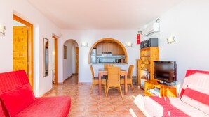 Ein Wohnzimmer mit Sofa, einem Esstisch mit Stühlen, einer Küchenzeile mit Fenster und einem Fernseher auf einem Lowboard.