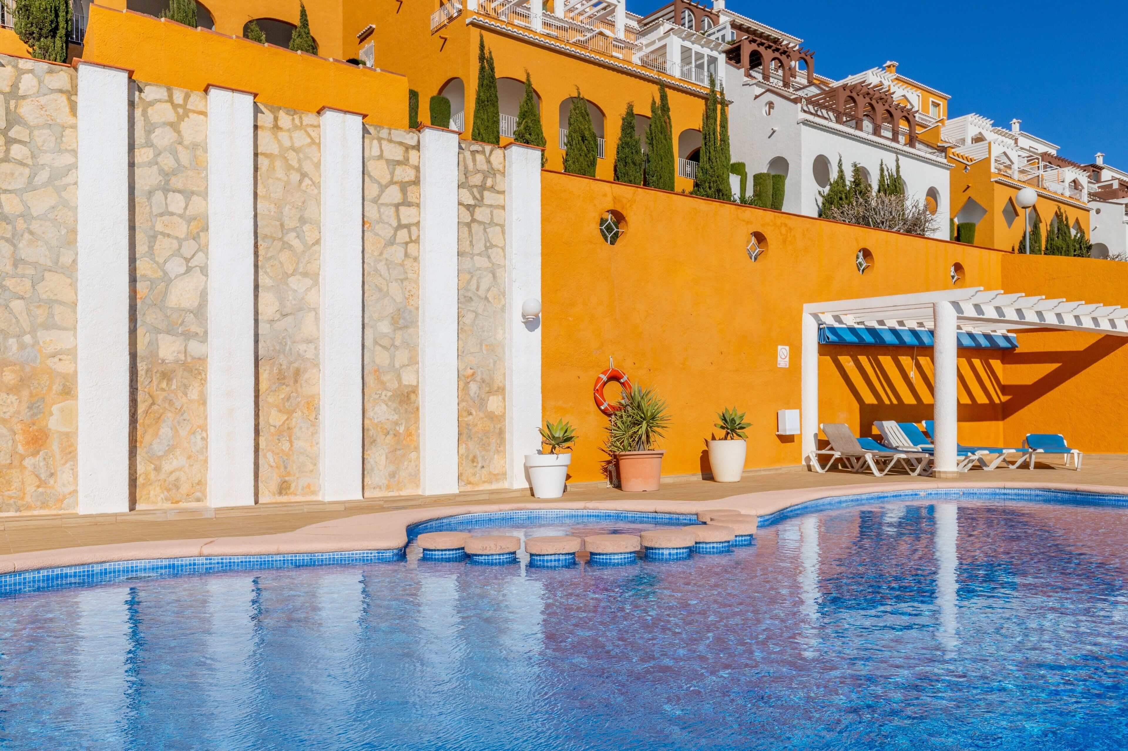 Una piscina con sillones de descanso y una pérgola, rodeada por un muro de piedra y un edificio amarillo con arcos.