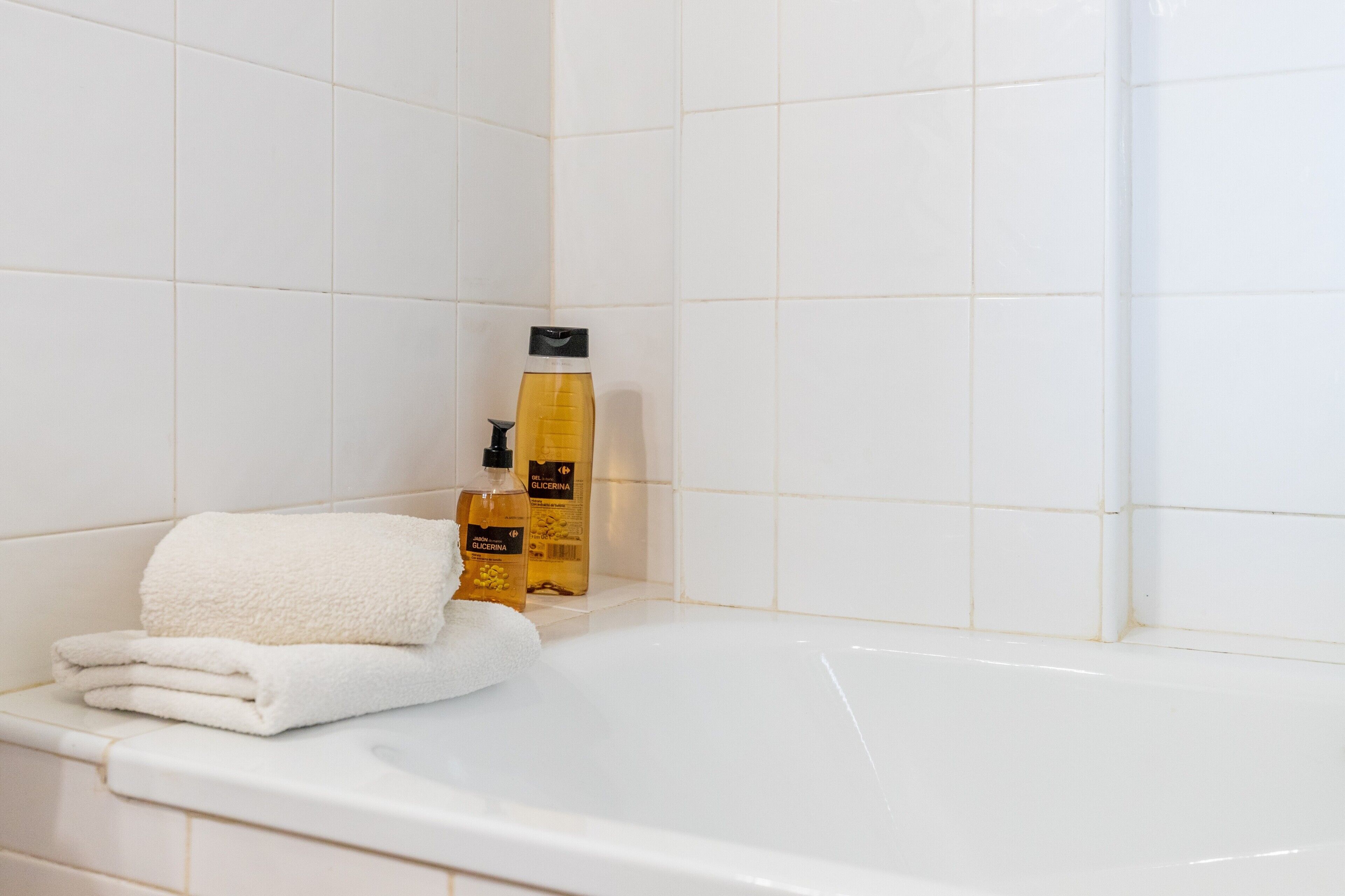 A white tiled bathroom with a white bathtub, a folded white towel, and a bottle of lotion.
