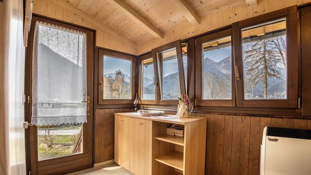 Interior de una cabaña de madera con cocina, ventanas amplias con vistas a la montaña y techo de madera.