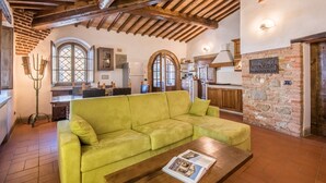 Un salon chaleureux avec un canapé vert, des poutres en bois, un mur en briques et une cuisine en arrière-plan.