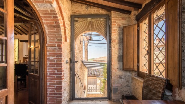 Un couloir étroit avec une porte en bois donnant sur une cour, des murs en pierre et une chaise en osier.