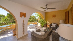 Un patio techado con mobiliario de mimbre, ventilador de techo y vista a la vegetación.