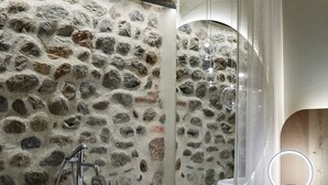 Une salle de bain avec un mur en pierre, une baignoire, un miroir avec un éclairage intégré et un lit recouvert d’une couette blanche.