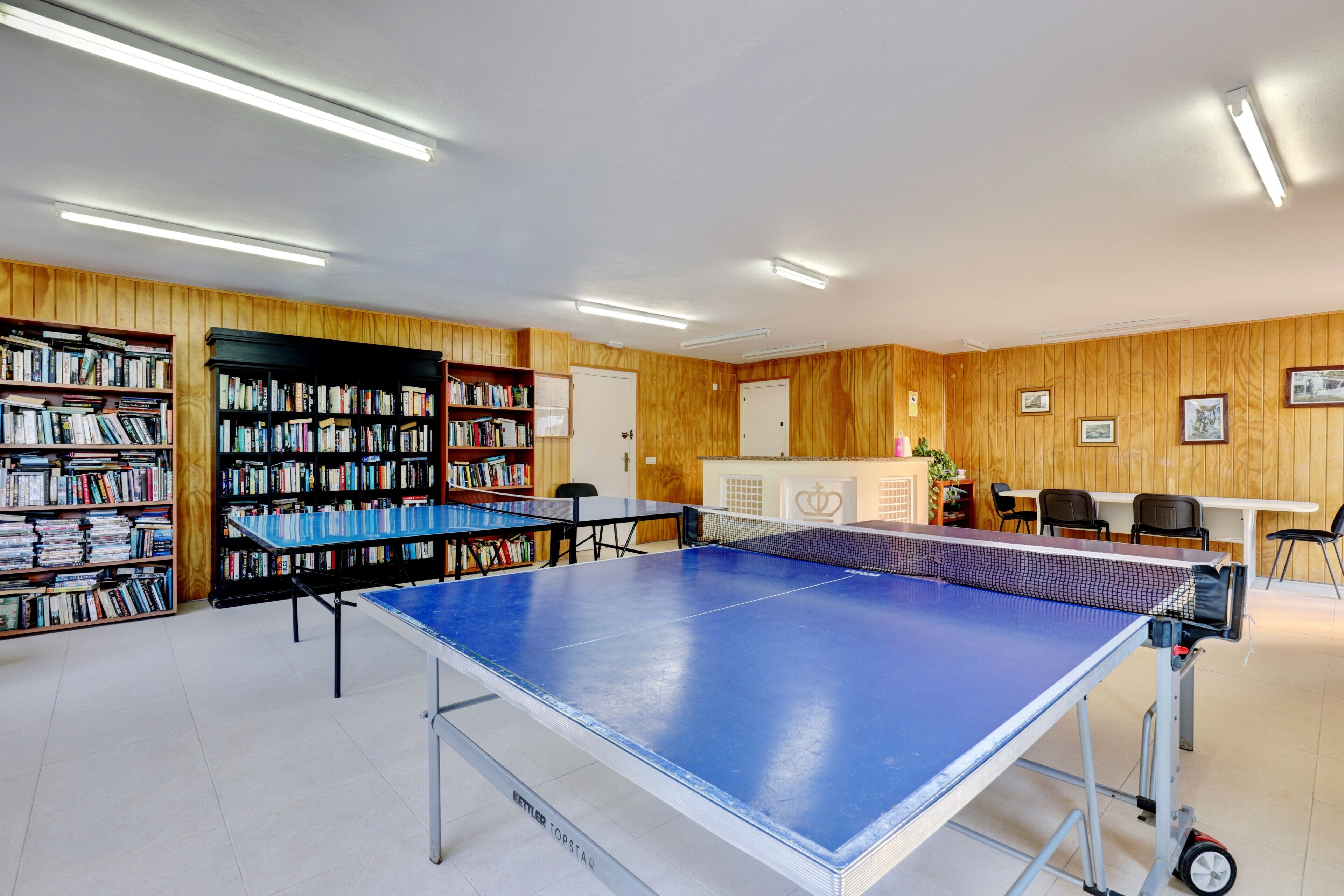 Een kamer met een tafeltennistafel, boekenplanken, een bureau, stoelen en een deur.