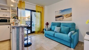 Una sala de estar con un sofá azul verdoso, una zona de comedor con una mesa y sillas, una cocina con microondas, y un balcón con cortinas amarillas.