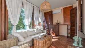 Una stanza con un divano, un tavolino in vimini, una lampada pendente in vimini e vista su del verde all'esterno.