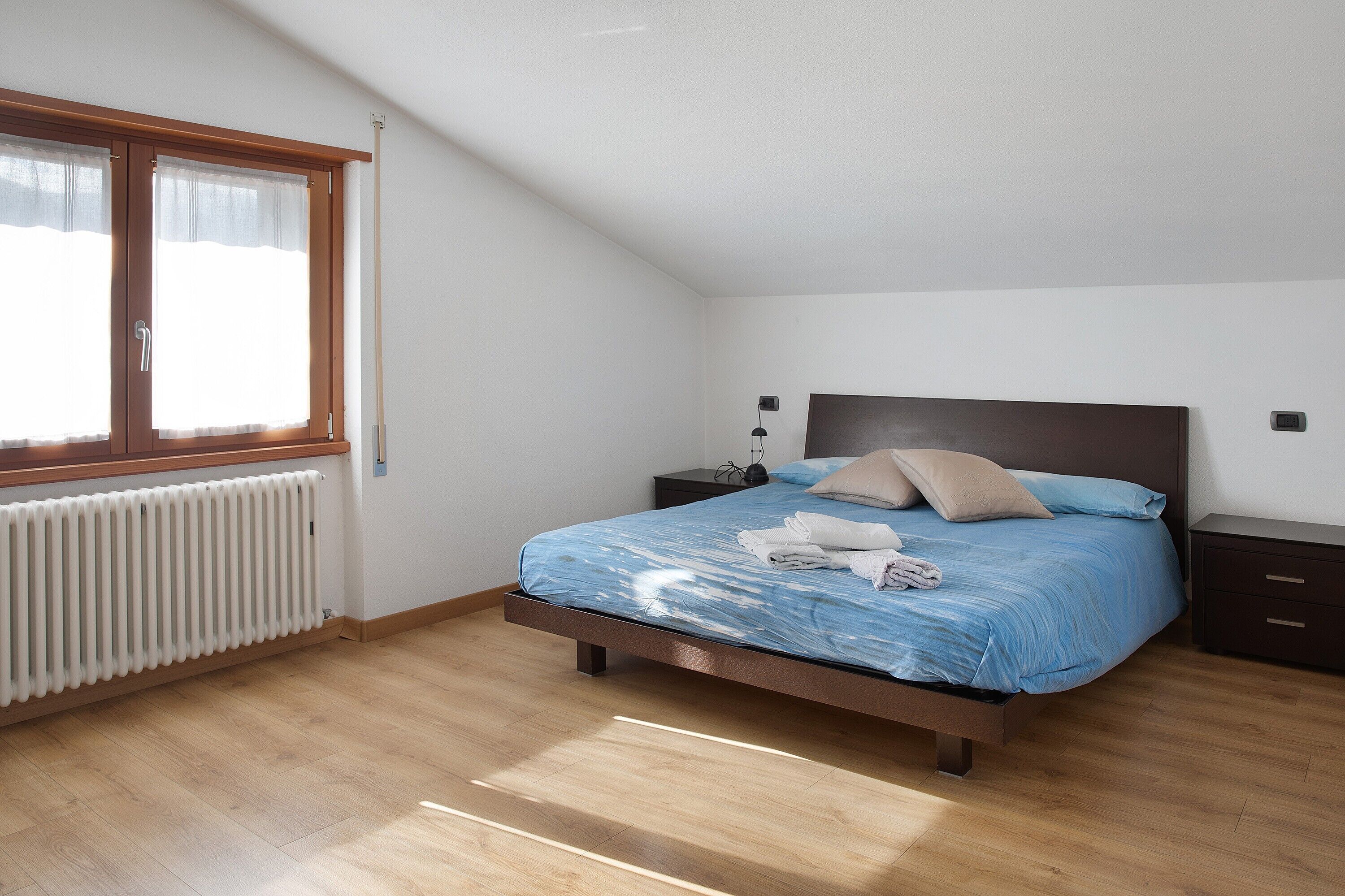 A bedroom with a bed, a nightstand, a lamp, a window with curtains, and a radiator.
