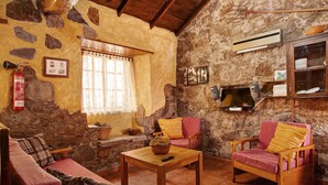 Un intérieur rustique avec des murs en pierre, un plafond en bois, une cheminée, un canapé, un fauteuil, une table et un téléviseur.