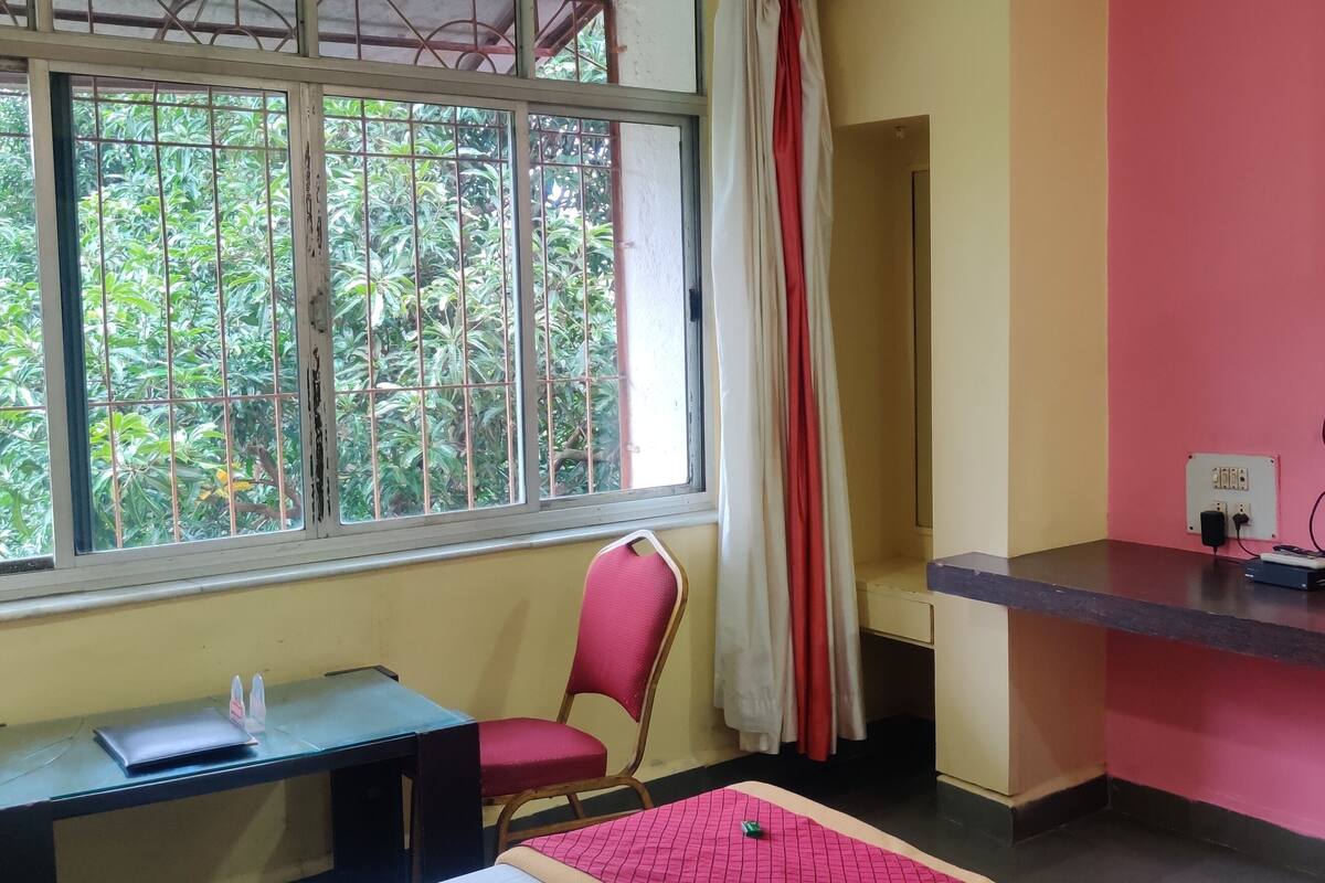 A room with a desk, chair, TV, and window with a view of greenery.