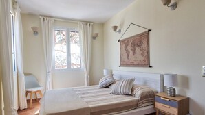 Un dormitorio con cama, mesita de noche, silla, ventana con cortinas y un mapa colgado en la pared.