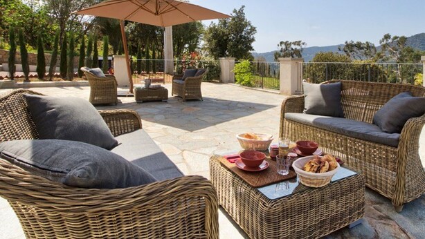 Un patio exterior con mobiliario de mimbre, una mesa con pan y bebidas, y vistas a árboles y colinas.